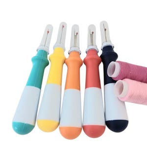 May include: Five seam rippers with ergonomic handles in teal, yellow, orange, red, and dark blue, each featuring a clear plastic cap and a metal blade. Two spools of thread in shades of pink are positioned horizontally across the dark blue tool.