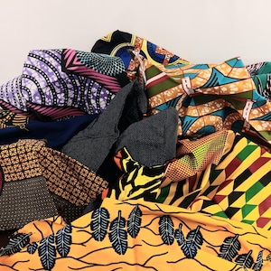 May include: A pile of colorful African wax print fabrics. The fabrics feature a variety of patterns, including geometric shapes, floral designs, and abstract motifs. Colors include yellow, orange, blue, black, and green.