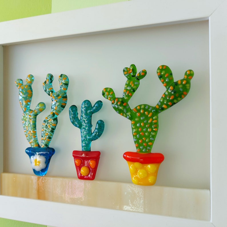 K&ouml;nnte beinhalten: Drei Glaskaktussculpturen in bunten T&ouml;pfen. Die Kakteen sind gr&uuml;n mit orangefarbenen und wei&szlig;en Akzenten. Die T&ouml;pfe sind rot, gelb und blau.