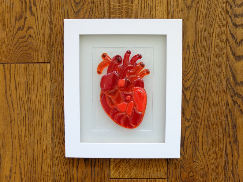 May include: A framed glass art piece depicting a red anatomical heart. The heart is made of stained glass and is set against a white background.