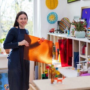 May include: A woman in a blue apron holds a sheet of orange textured glass in a workshop. The workshop has shelves filled with colorful glass art and supplies.