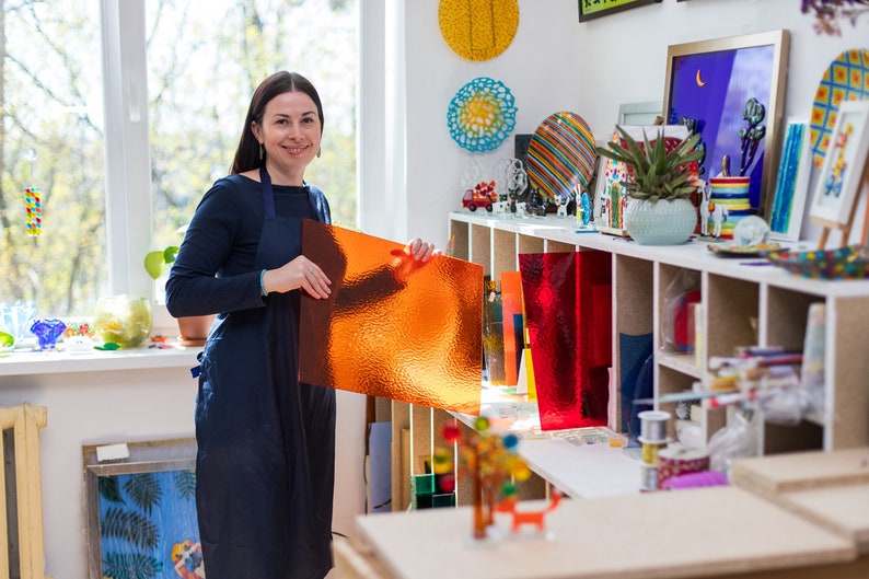May include: A woman in a blue apron holds a sheet of orange textured glass in a workshop. The workshop has shelves filled with colorful glass art and supplies.