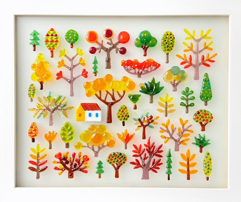 May include: A framed artwork featuring a collection of colorful glass trees and a small house. The trees are in various shapes and sizes, with vibrant hues of red, orange, yellow, and green. The house has a red roof and yellow walls.