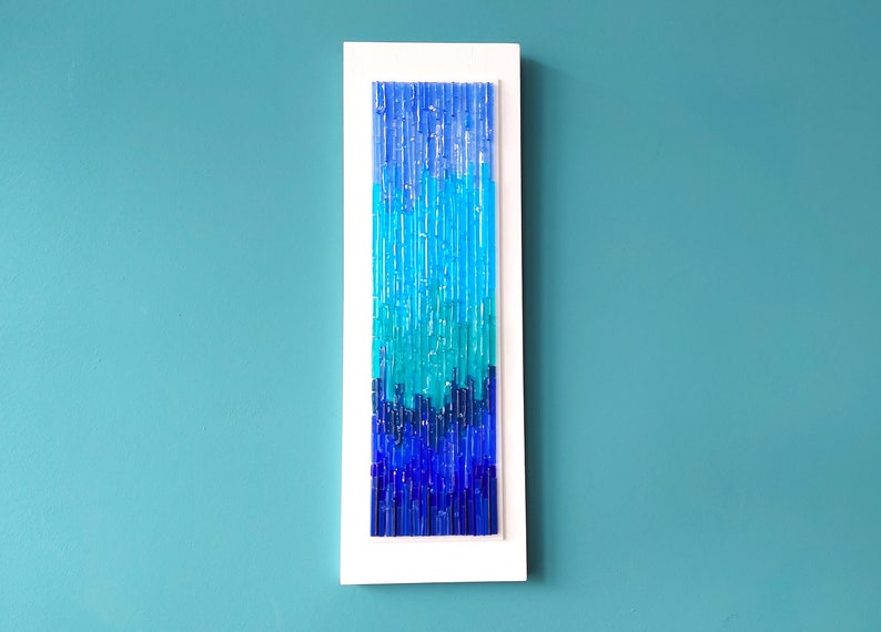 May include: A vertical wall art piece featuring a gradient of blue glass rods. The rods transition from light turquoise to deep blue, encased in a white rectangular frame. The artwork is mounted on a teal wall.