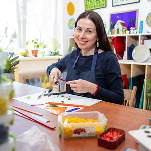May include: A woman works on a stained glass project in her studio. She is using a glass cutter to cut a piece of glass. There are various tools and materials on the table, including rulers, glass cutters, and small containers of colored glass tiles.