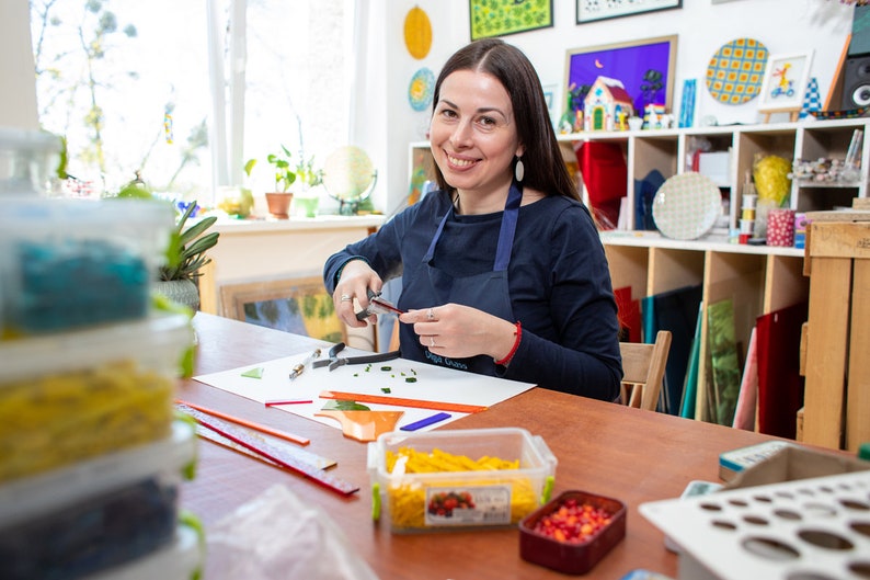 May include: A woman works on a stained glass project in her studio. She is using a glass cutter to cut a piece of glass. There are various tools and materials on the table, including rulers, glass cutters, and small containers of colored glass tiles.