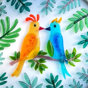 May include: Two glass birds, one yellow with orange spots and one blue with green accents, are perched on a branch with green leaves. The birds are facing each other and appear to be kissing.