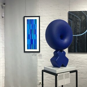 May include: A blue abstract sculpture with a round top and a base shaped like an X. The sculpture is on a white pedestal with a black base. There are three paintings on the wall behind the sculpture. The paintings are all abstract and feature blue and black colors.
