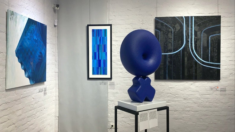 May include: A blue abstract sculpture with a round top and a base shaped like an X. The sculpture is on a white pedestal with a black base. There are three paintings on the wall behind the sculpture. The paintings are all abstract and feature blue and black colors.