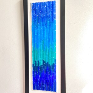 May include: Abstract wall art in a black frame. The artwork features a vertical arrangement of blue glass rods, transitioning from dark blue at the bottom to lighter shades at the top. The piece is mounted on a white background.