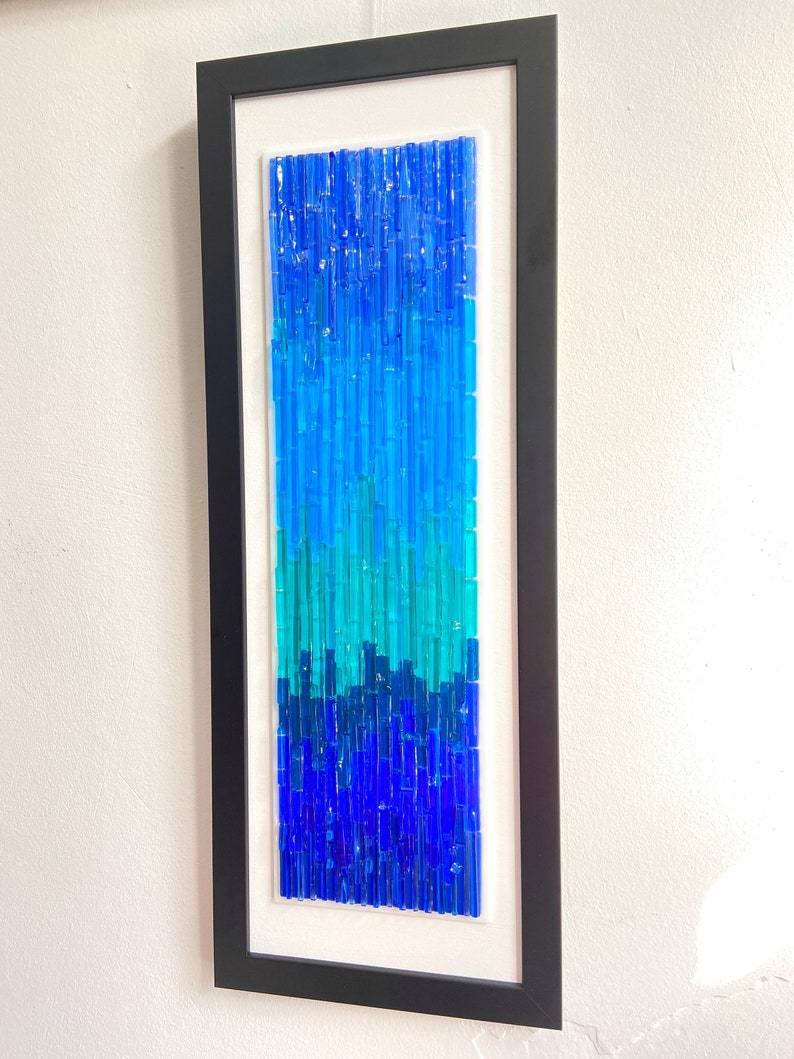 May include: Abstract wall art in a black frame. The artwork features a vertical arrangement of blue glass rods, transitioning from dark blue at the bottom to lighter shades at the top. The piece is mounted on a white background.