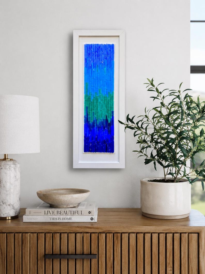 May include: A framed abstract art piece with a vertical design. The artwork features a gradient of blues and greens, encased in a white rectangular frame. The piece is displayed on a wooden cabinet, alongside decorative items and a potted plant.