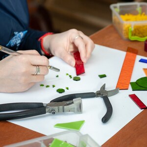 May include: A person working on a mosaic project with glass tiles, tools, and rulers on a white surface. The tools include a pair of pliers, a glass cutter, and a ruler. The glass tiles are in various colors, including green, red, and orange.