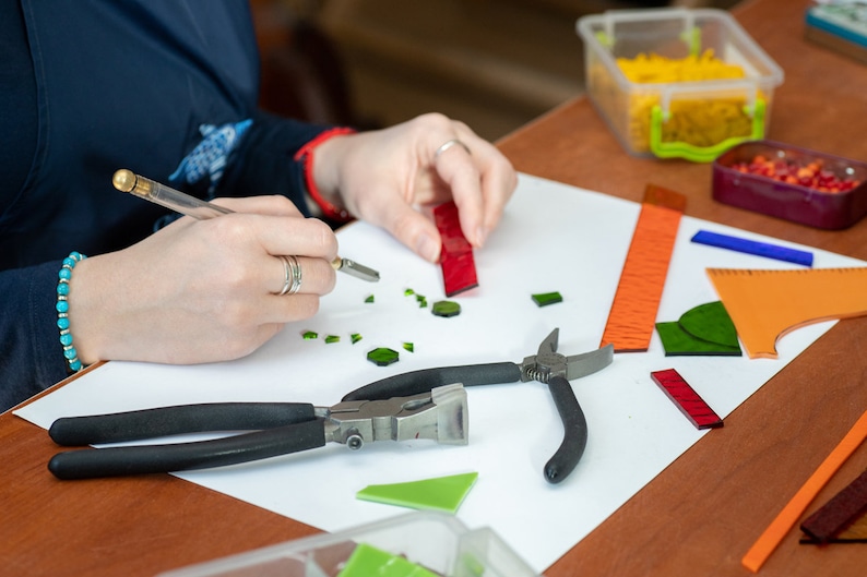 May include: A person working on a mosaic project with glass tiles, tools, and rulers on a white surface. The tools include a pair of pliers, a glass cutter, and a ruler. The glass tiles are in various colors, including green, red, and orange.