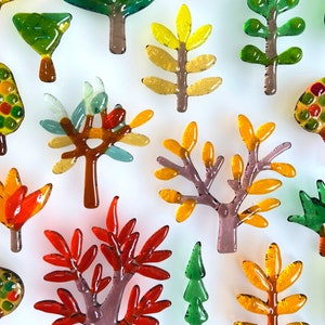 May include: A collection of colorful glass tree ornaments in various shapes and sizes. The trees feature shades of green, yellow, orange, and red, with brown and purple trunks. The ornaments are arranged against a white background, creating a vibrant display.