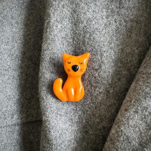 May include: A glossy orange cat-shaped brooch. The cat has a black nose and eyes, and is set against a grey fabric background. The cat is sitting with its tail curled.