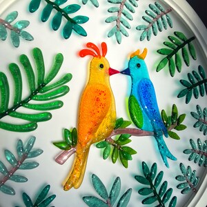 May include: A white circular framed wall art piece with a yellow and blue glass bird design. The birds are perched on a branch with green glass leaves surrounding them.