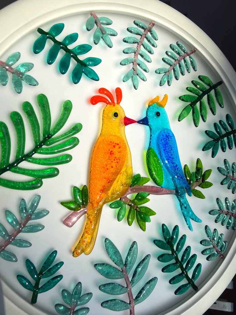 May include: A white circular framed wall art piece with a yellow and blue glass bird design. The birds are perched on a branch with green glass leaves surrounding them.
