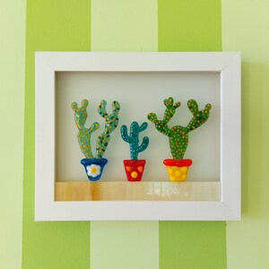 K&ouml;nnte beinhalten: Drei Glaskakteen in bunten T&ouml;pfen, gerahmt in einem wei&szlig;en Rahmen. Die Kakteen sind gr&uuml;n mit gelben und orangefarbenen Akzenten. Die T&ouml;pfe sind blau, rot und gelb.