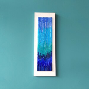 May include: A rectangular wall art piece with a white frame. The artwork features a gradient of blue hues, from light turquoise to deep indigo, created with vertical glass rods. The piece is mounted on a teal wall.