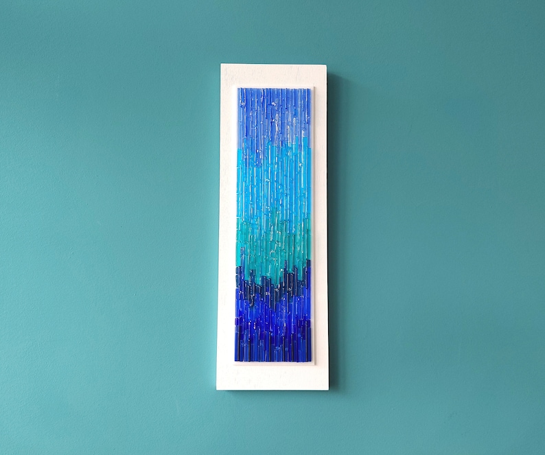 May include: A rectangular wall art piece with a white frame. The artwork features a gradient of blue hues, from light turquoise to deep indigo, created with vertical glass rods. The piece is mounted on a teal wall.