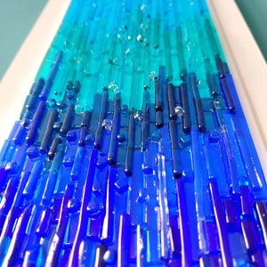 May include: Abstract art piece featuring a gradient of blues and teals. Vertical glass rods create a textured surface, transitioning from deep blue at the base to lighter shades at the top. Clear glass accents add dimension.