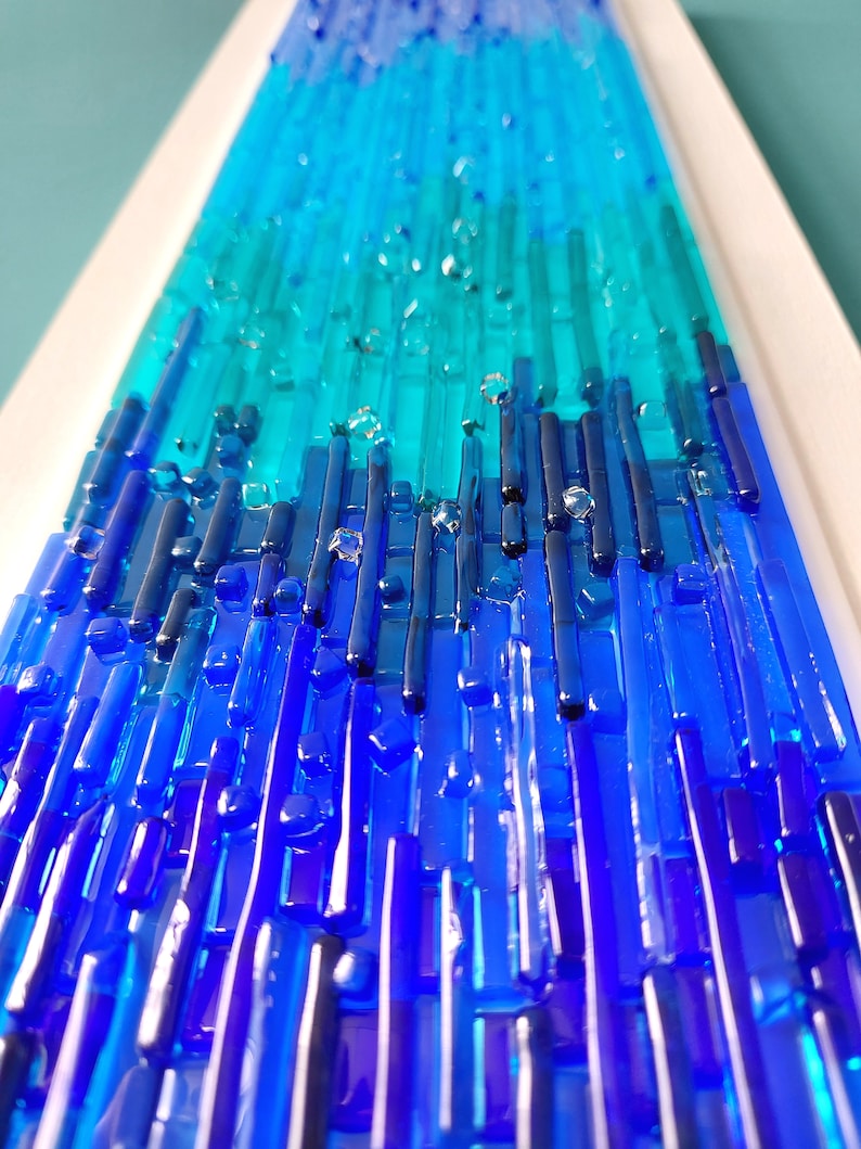May include: Abstract art piece featuring a gradient of blues and teals. Vertical glass rods create a textured surface, transitioning from deep blue at the base to lighter shades at the top. Clear glass accents add dimension.