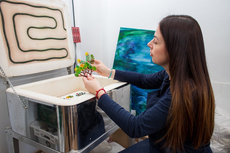 May include: A woman holds a glass art piece shaped like a tree with green and yellow glass leaves. The piece is in front of a kiln with the door open.