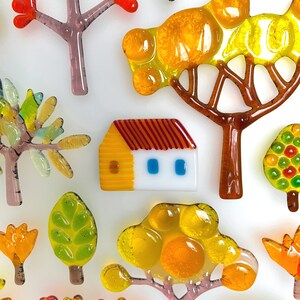 May include: A collection of colorful glass art pieces depicting trees, leaves, and a small house. The pieces feature vibrant hues of orange, yellow, green, and red, with brown and purple accents. The art pieces are arranged on a white background.