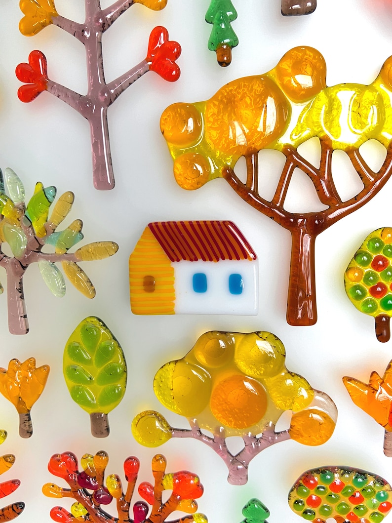 May include: A collection of colorful glass art pieces depicting trees, leaves, and a small house. The pieces feature vibrant hues of orange, yellow, green, and red, with brown and purple accents. The art pieces are arranged on a white background.