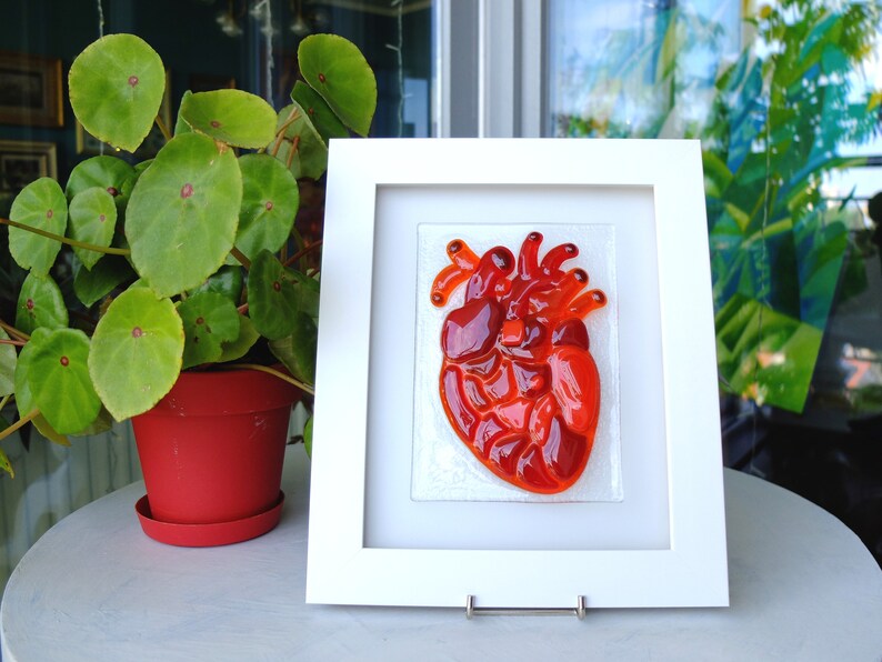 May include: A framed glass art piece depicting a red anatomical heart. The heart is made of multiple pieces of red glass, creating a textured and dimensional effect.