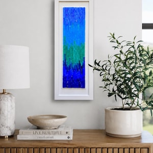May include: A framed abstract art piece with a vertical design. The artwork features a gradient of blues and greens, encased in a white rectangular frame. The piece is displayed on a wooden cabinet, alongside a lamp, books, and a decorative bowl.