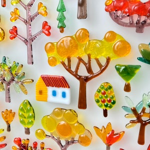 May include: A collection of colorful glass tree and house ornaments. The trees feature various designs with red, orange, yellow, and green leaves. A small house with a yellow facade and red roof is also included.