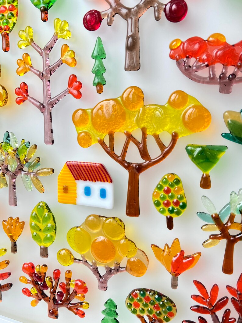 May include: A collection of colorful glass tree and house ornaments. The trees feature various designs with red, orange, yellow, and green leaves. A small house with a yellow facade and red roof is also included.