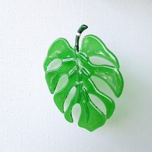 Könnte beinhalten: Eine leuchtend grüne Monstera-Blattdekoration aus Glas, die an einem dünnen weißen Faden hängt. Das Blatt weist charakteristische Fensterungen und eine glänzende Oberfläche auf und verleiht der Dekoration einen Hauch von Natur. Das Blatt ist etwa 15 cm lang.