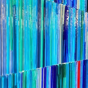 May include: A close-up of a wall art piece made of many thin glass rods in shades of blue, teal, and green. The rods are arranged in vertical lines, creating a textured and abstract pattern.
