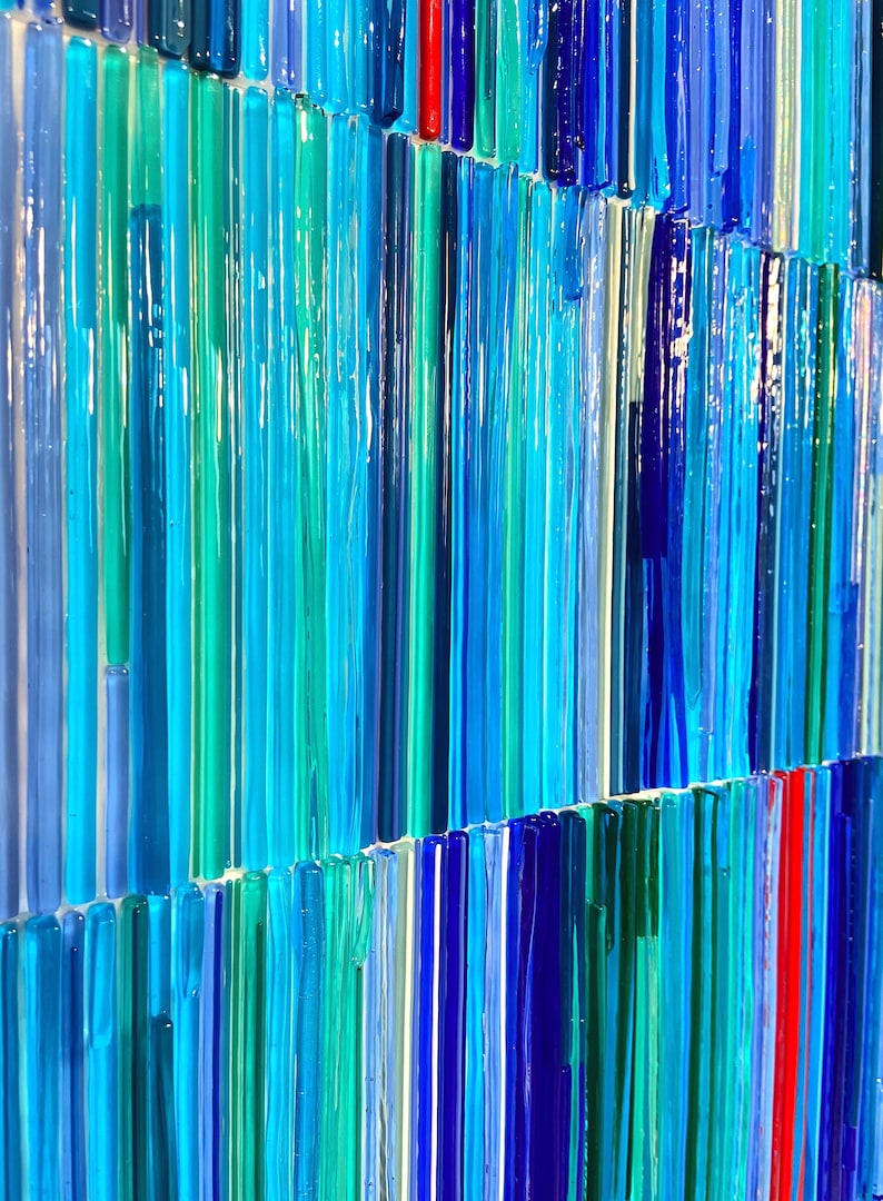 May include: A close-up of a wall art piece made of many thin glass rods in shades of blue, teal, and green. The rods are arranged in vertical lines, creating a textured and abstract pattern.