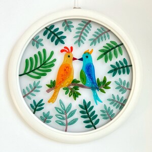 May include: A white circular frame with a glass panel featuring two colorful birds perched on a branch surrounded by green leaves.