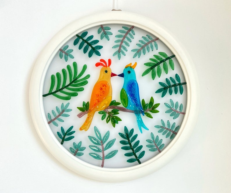 May include: A white circular frame with a glass panel featuring two colorful birds perched on a branch surrounded by green leaves.