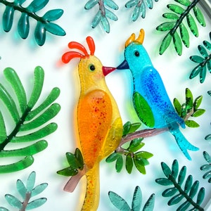 May include: Two colorful glass birds perched on a branch with green glass leaves. The bird on the left is yellow with orange accents and the bird on the right is blue with green accents.