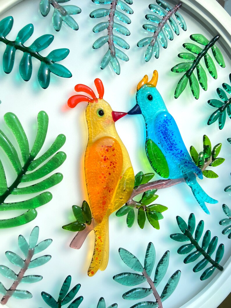 May include: Two colorful glass birds perched on a branch with green glass leaves. The bird on the left is yellow with orange accents and the bird on the right is blue with green accents.