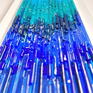 May include: Abstract glass art piece featuring a gradient of blues and teals. Vertical glass rods create a textured surface within a white frame. The artwork evokes a sense of depth and movement, resembling water or a stylized landscape.