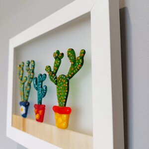 K&ouml;nnte beinhalten: Ein wei&szlig; gerahmtes Kunstwerk mit drei Glaskakteen in bunten T&ouml;pfen. Die Kakteen sind gr&uuml;n mit orangefarbenen und gelben Akzenten. Die T&ouml;pfe sind rot, blau und gelb.