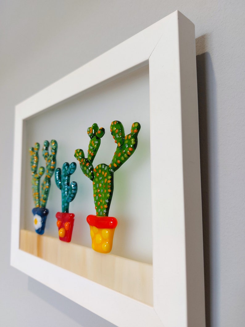 K&ouml;nnte beinhalten: Ein wei&szlig; gerahmtes Kunstwerk mit drei Glaskakteen in bunten T&ouml;pfen. Die Kakteen sind gr&uuml;n mit orangefarbenen und gelben Akzenten. Die T&ouml;pfe sind rot, blau und gelb.