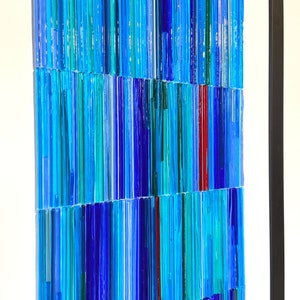 May include: A framed artwork made of colorful glass rods in shades of blue, teal, and red. The rods are arranged in a vertical pattern, creating a textured and abstract design.