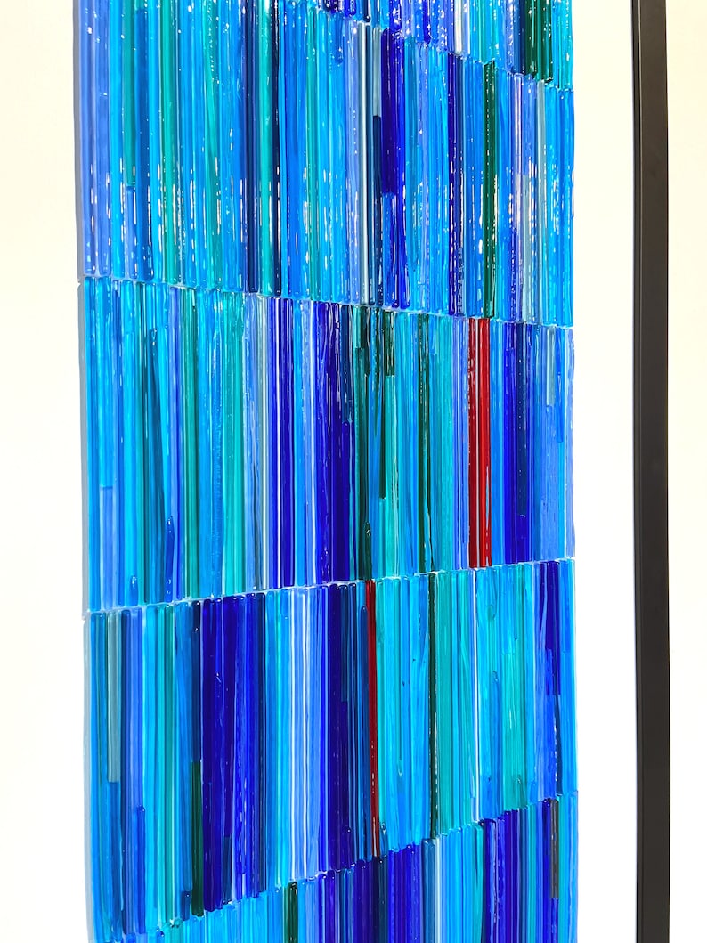 May include: A framed artwork made of colorful glass rods in shades of blue, teal, and red. The rods are arranged in a vertical pattern, creating a textured and abstract design.