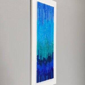 May include: Vertical abstract wall art with a white frame. The artwork features a gradient of blue and teal glass rods, creating a textured, modern design. The color palette transitions from deep blue at the bottom to lighter shades at the top.