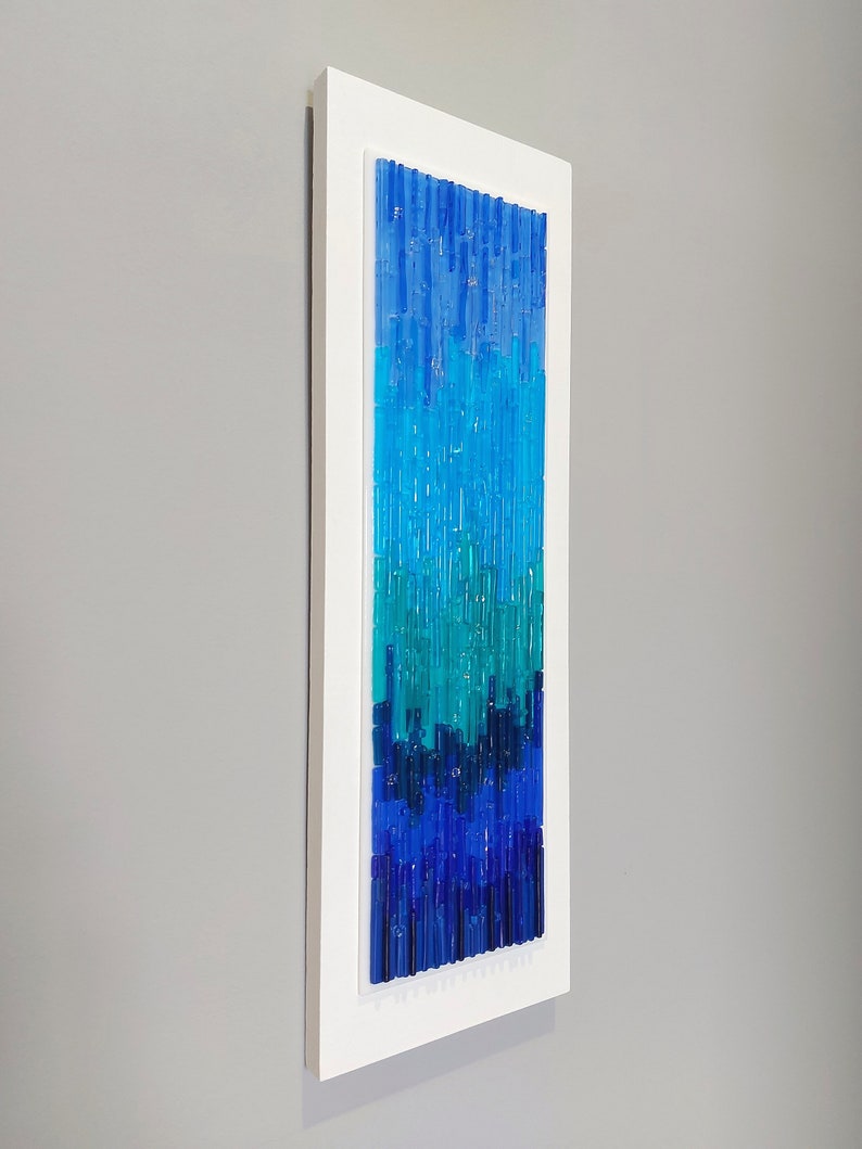 May include: Vertical abstract wall art with a white frame. The artwork features a gradient of blue and teal glass rods, creating a textured, modern design. The color palette transitions from deep blue at the bottom to lighter shades at the top.