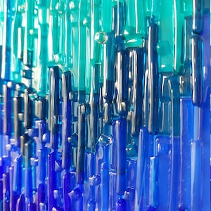 May include: Abstract art piece featuring vertical glass rods in shades of blue and teal. The rods vary in length and thickness, creating a textured, layered effect. The color gradient transitions from light teal at the top to deep blue at the bottom.