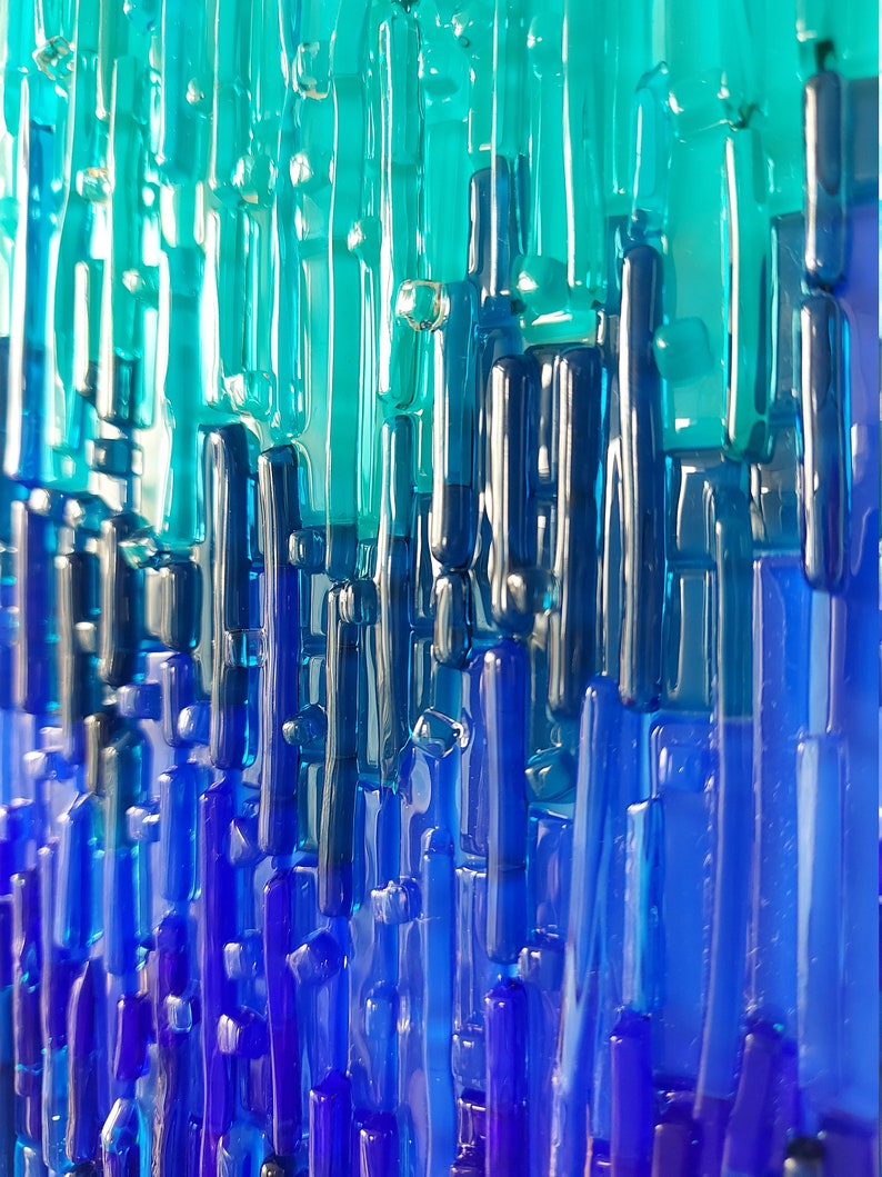 May include: Abstract art piece featuring vertical glass rods in shades of blue and teal. The rods vary in length and thickness, creating a textured, layered effect. The color gradient transitions from light teal at the top to deep blue at the bottom.